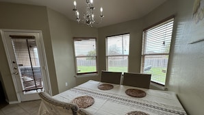Une salle à manger avec une table dressée pour quatre personnes, un lustre et une vue sur le jardin à travers de grandes fenêtres.
