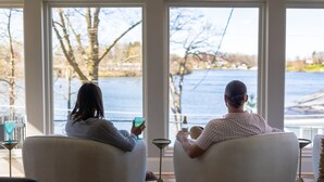 Two people seated by large windows overlooking a body of water, with a table and a lamp between them. - Hilltop Lakehouse - Free Kayaks, Deck, Hot Tub , Pet Friendly (Akron)