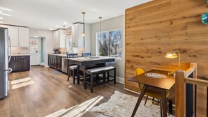 A modern kitchen with wooden flooring, a dining area with a wooden table and chairs, and a large window providing natural light. - Hilltop Lakehouse - Free Kayaks, Deck, Hot Tub , Pet Friendly (Akron)