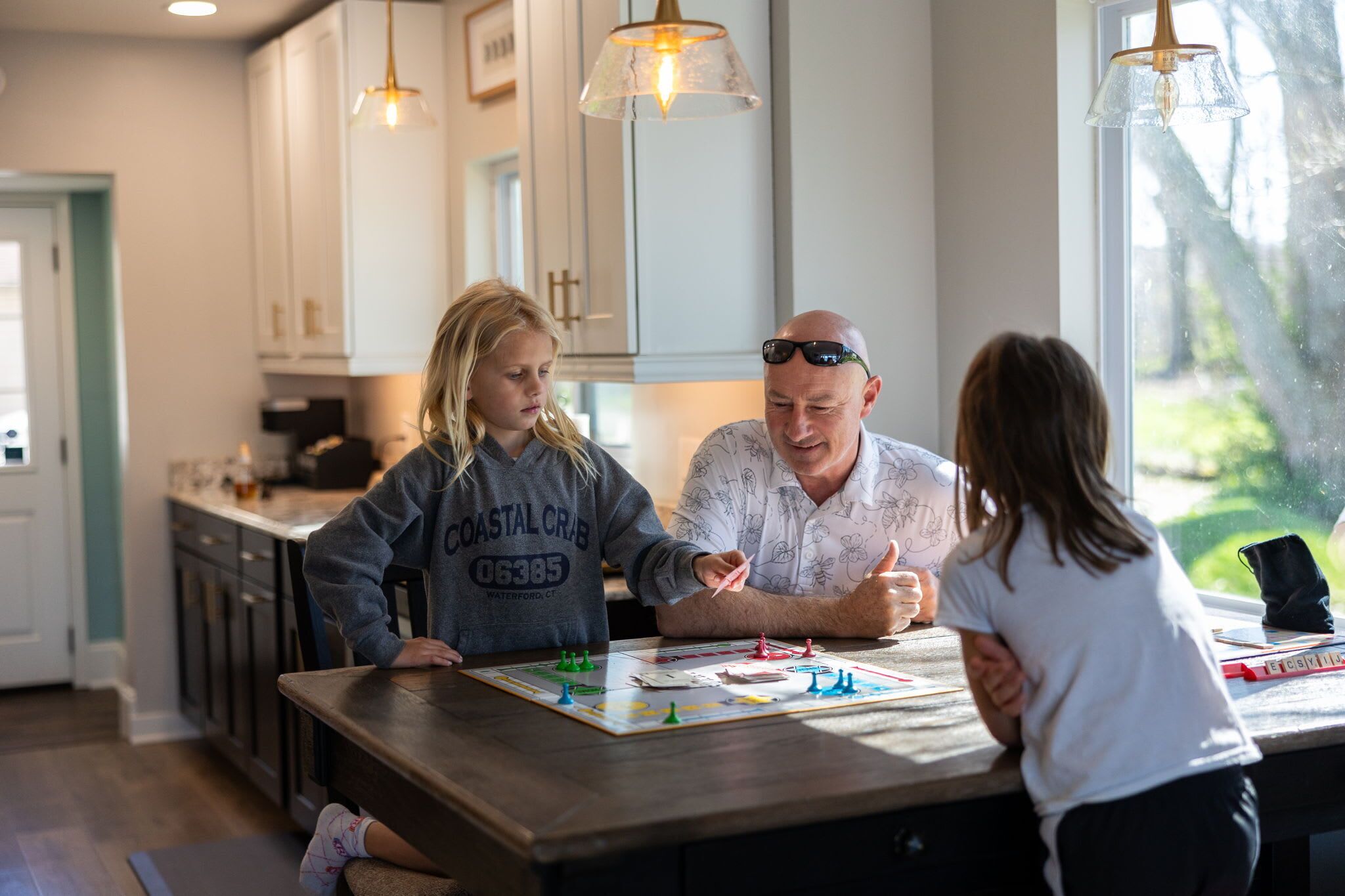 A family playing a board game at a kitchen island with white cabinets and pendant lights.