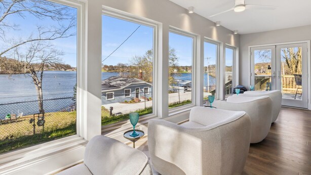 A spacious room with large windows offering a view of a lake and a house. There are white chairs and a small table with a decorative item. - Hilltop Lakehouse - Free Kayaks, Deck, Hot Tub , Pet Friendly (Akron)