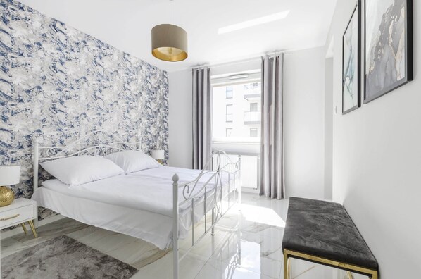 Ein modernes Schlafzimmer mit einem weißen Bett, einer Wand mit Marmoroptik, einem Nachttisch mit Lampe und Blick auf ein Gebäude durch ein Fenster.