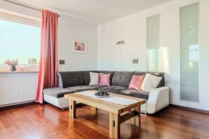 Un salon moderne avec un canapé gris, une table basse en bois et une grande fenêtre agrémentée d’un rideau rose.