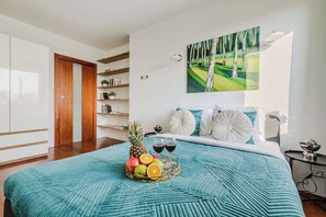 A bedroom with a bed, a fruit basket, two wine glasses, and a painting on the wall.