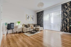 A modern living room with a sofa, coffee table, and a desk with a chair. There are two framed pictures on the wall, a vase with flowers, and a large window with curtains.