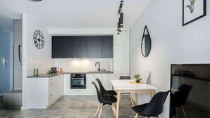 A modern kitchen with dark cabinets, a white countertop, and a dining area with a table and chairs. - Lumina airy apart near Manufaktura (Łódź)