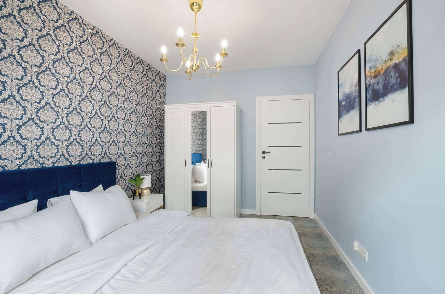 Een slaapkamer met behang met patroon, een bed met witte lakens, een kroonluchter, een kledingkast en twee ingelijste foto's aan de muur.