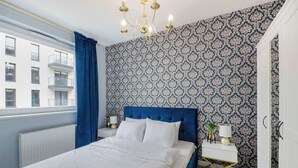A bedroom with a patterned wallpaper, a blue headboard, a white bed, a white nightstand, a gold lamp, and a view of a building outside. - Lumina airy apart near Manufaktura (Łódź)