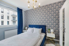 Une chambre avec un papier peint à motifs, une tête de lit bleue, un lit blanc, une table de chevet blanche, une lampe dorée et une vue sur un bâtiment à l’extérieur.