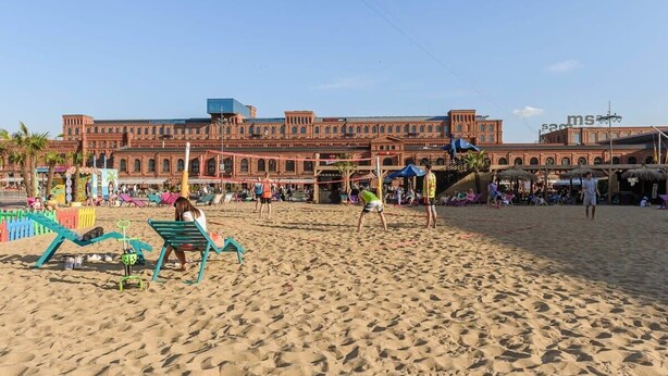 Une plage de sable avec un grand bâtiment en arrière-plan, un filet de volley-ball et des personnes qui jouent au volley-ball.