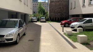 Parkkialue, jossa autoja on pysäköitynä molemmille puolille, asfaltoitu kävelyreitti ja taustalla näkyviä rakennuksia.