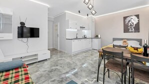 Moderni hotellihuone, jossa on seinälle asennettu taulutelevisio, ruokailutila pöydän ja tuolien kera, valkoisilla kaapeilla varustettu keittiö ja sänky, jonka päällä on pehmolelu.