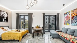 Moderni hotellihuone, jossa on sänky, sohva, ruokapöytä ja suuret ikkunat, joista avautuu näkymä kaupunkiin.