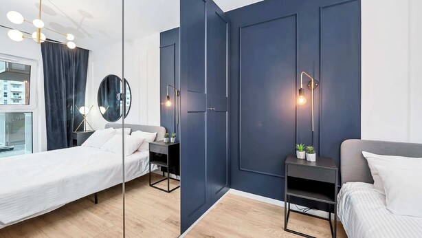 Un dormitorio moderno con un espejo grande, dos camas, mesitas de noche con lámparas y vistas a la ciudad a través de una ventana.