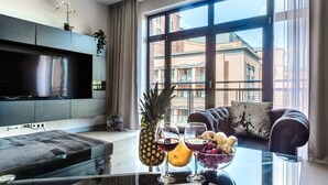 Een moderne woonkamer met een groot raam, een flatscreen televisie en een glazen salontafel met fruit en glazen.