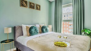 Un dormitorio con cama, una mesita de noche, una lámpara, una ventana con cortinas y vistas a edificios.