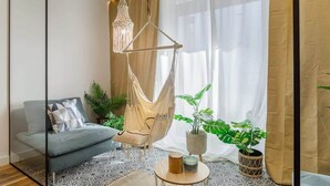 Een moderne woonkamer met een grijze bank, een hangstoel van macramé, een ronde salontafel en een groot raam met lichte gordijnen.