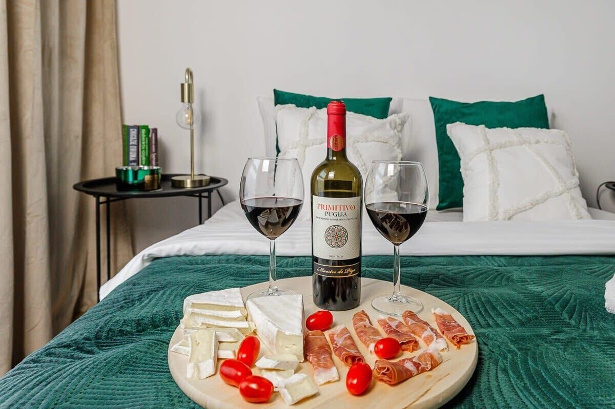 A bed with a green blanket, a wine bottle, two glasses of red wine, a cheese platter with various cheeses and tomatoes, and a small table with books and a lamp in the background.
