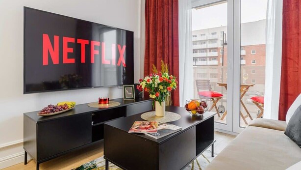 Una sala de estar moderna con un televisor grande mostrando el logotipo de Netflix, un mueble de entretenimiento negro, un florero con flores y un balcón con cortinas rojas.