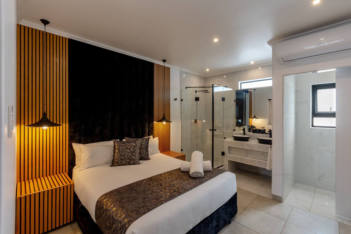 Quarto de hotel moderno com uma cama grande, box de vidro para o chuveiro, toucador com espelho e vista para a cidade.