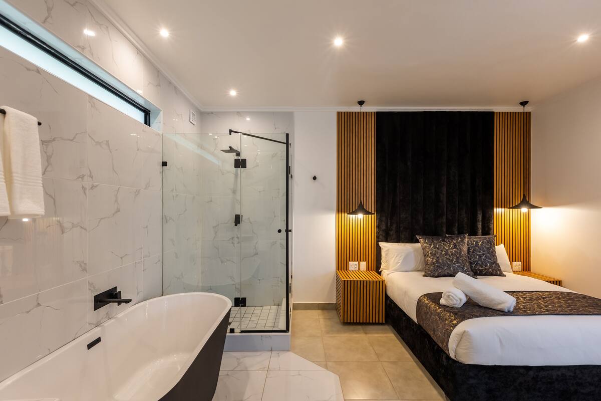 Quarto moderno com cama grande, mesas de cabeceira, um criado-mudo, chuveiro com box de vidro e banheiro com banheira e paredes revestidas de mármore.