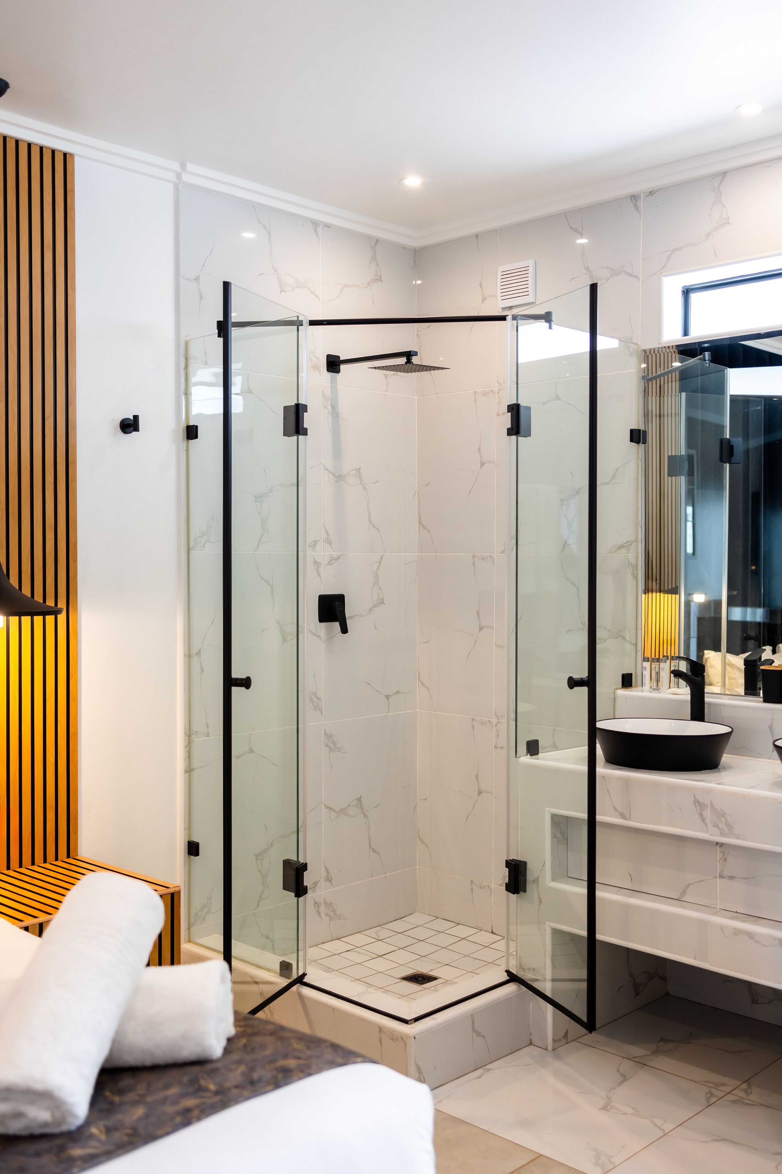 A modern bathroom with a glass-enclosed shower, marble tiles, a wooden bench, and a large mirror.