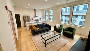 Ein geräumiger Wohnbereich mit einer Couch, einem Couchtisch und einem grünen Sessel. Im Hintergrund ist eine Küche mit modernem Design zu sehen.