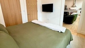 Ein Schlafzimmer mit einem großen Bett, einem flachbildfernseher an der Wand und Blick in den angrenzenden Küchenbereich.