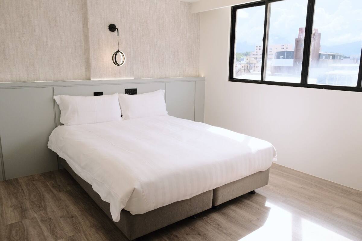 Un dormitorio moderno con cama, una lámpara fijada a la pared y ventana con vistas a la ciudad.