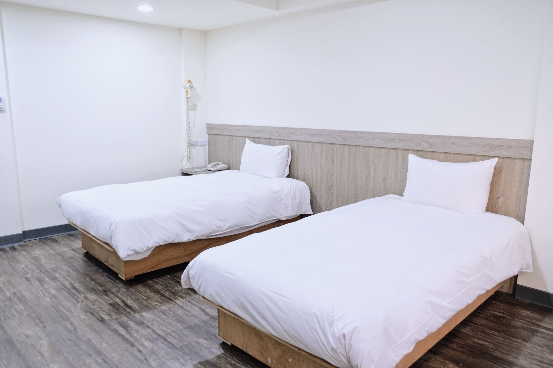 A hotel room with two single beds, wooden headboards, a bedside table with a lamp, and a phone.
