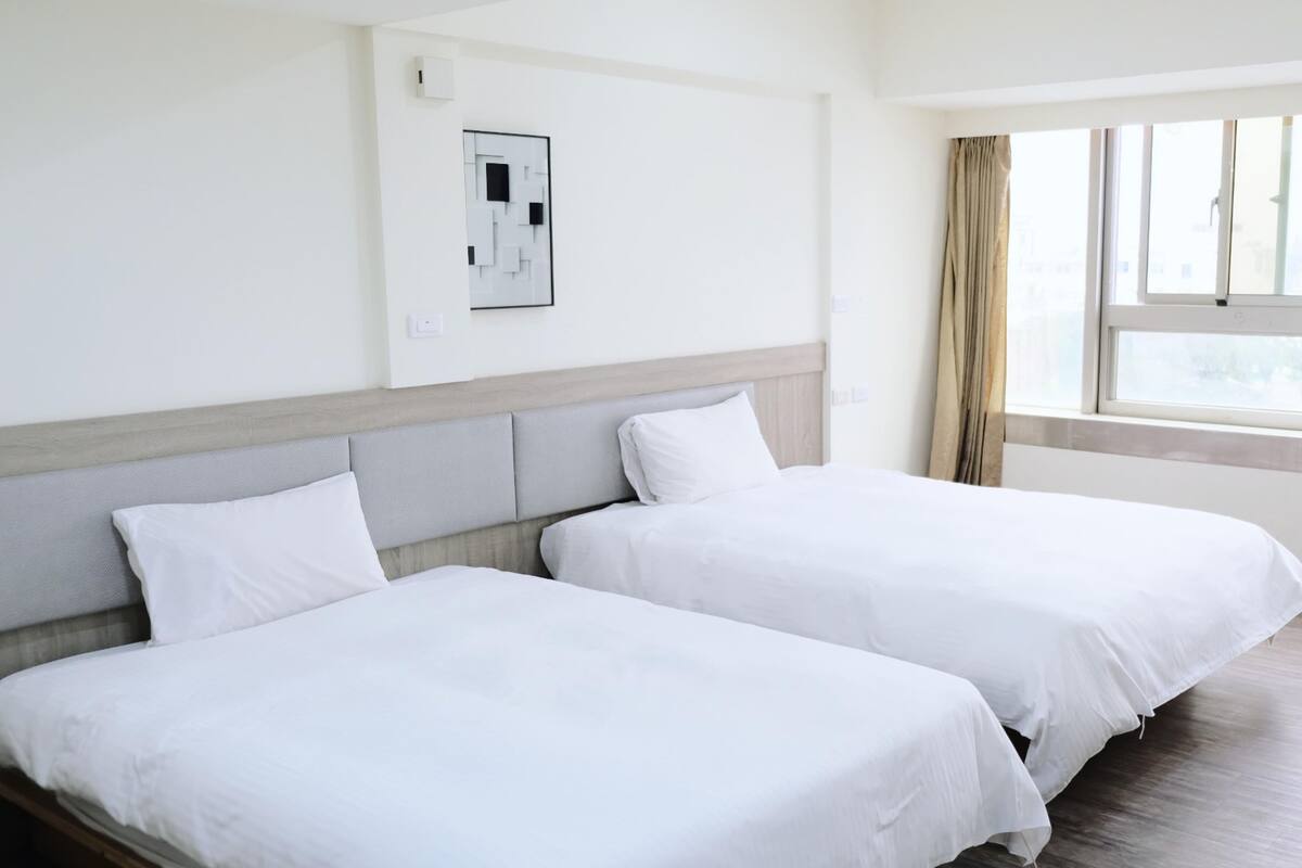 Habitación de hotel con dos camas individuales, cabecero de madera, ventana con cortinas y un interruptor de luz en la pared.
