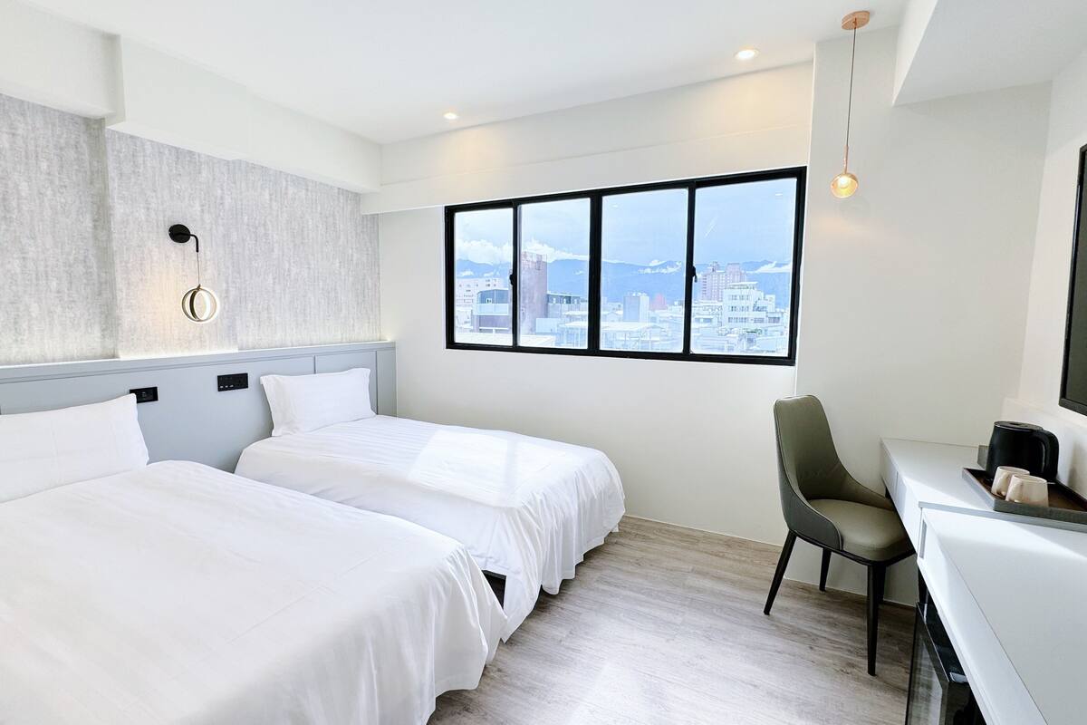 Habitación de hotel con dos camas, un escritorio, una silla, un ventanal grande con vista a la ciudad y una lámpara moderna.