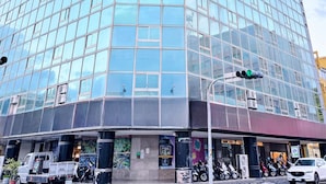 Een modern glazen gebouw met een hoingang, een parkeerplaats met motorfietsen en auto's, en een verkeerslicht.