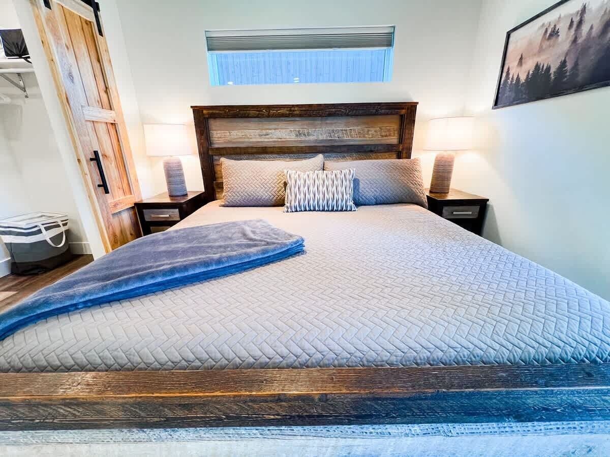 A large bed with a wooden headboard, two bedside lamps, a window with blinds, and a framed picture on the wall.