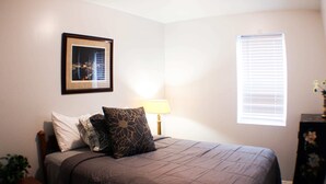A bedroom with a bed, a nightstand, a lamp, a window with blinds, and a framed picture on the wall. - Carey Way Quarters – Modern 2BR Near Carson Street (Pittsburgh)