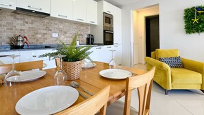 Eine moderne Küche mit weißen Schränken, einer Ziegelrückwand und Edelstahlgeräten. Ein Esstisch ist gedeckt mit Tellern, Besteck und einem Blumentopf. Im Wohnbereich sind ein gelber Sessel und eine Wanddekoration zu sehen.