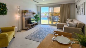 Ein modernes Wohnzimmer mit Fernseher, einer Couch, einem Esstisch mit Weinglas und Blick auf einen Pool und Grünflächen.