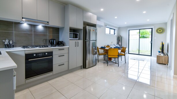 A modern kitchen with stainless steel appliances, a dining area with yellow chairs, and a large window. - Royal Lake Guest House (Toukley)