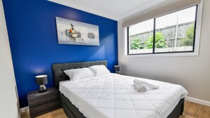 A bedroom with a large bed, a nightstand, a wall-mounted lamp, and a window with a view of greenery. - Royal Lake Guest House (Toukley)