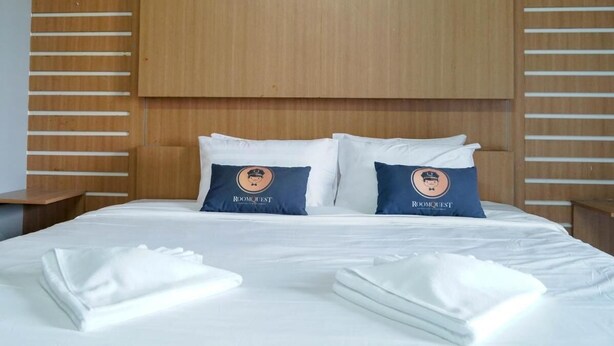 Et hotelværelse med en stor seng, to puder med 'RoomQuest'-branding og pænt foldede hvide håndklæder.
