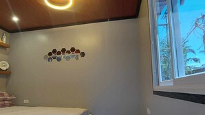 A bedroom with a bed, a wall-mounted shelf with decorative items, a window with a view of a palm tree and a power line structure. - B&F Travellers Inn (Daanbantayan)