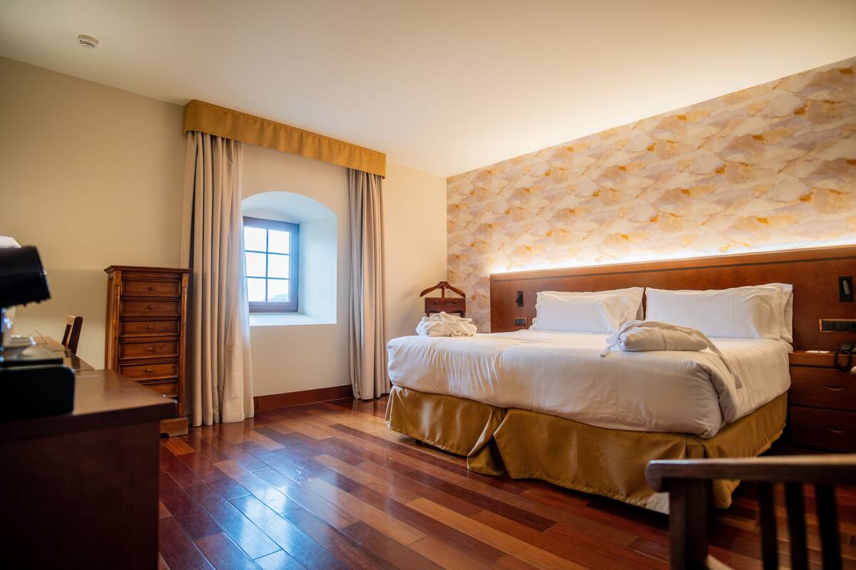 Quarto de hotel com piso de madeira, cama com roupa de cama branca, cômoda de madeira, escrivaninha com luminária e janela com cortinas.