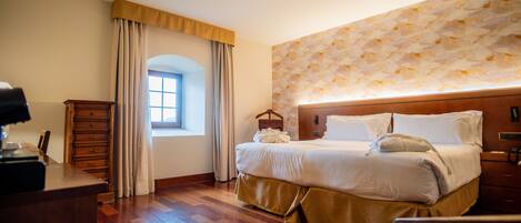 A hotel room with a wooden floor, a bed with white bedding, a wooden dresser, a desk with a lamp, and a window with curtains.