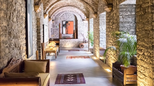 A stone-walled corridor with arches, a wooden bench, a table with a lamp, and a potted plant.