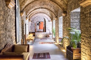 Un pasillo de paredes de piedra con arcos, un banco de madera, una mesa con una lámpara y una planta en maceta.