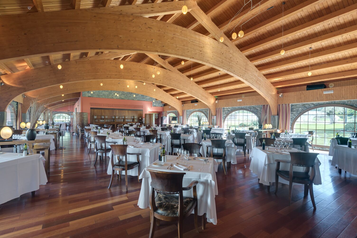 Uma sala de jantar ampla, com altos tetos de madeira e grandes janelas. Mesas dispostas com toalhas brancas e cadeiras.