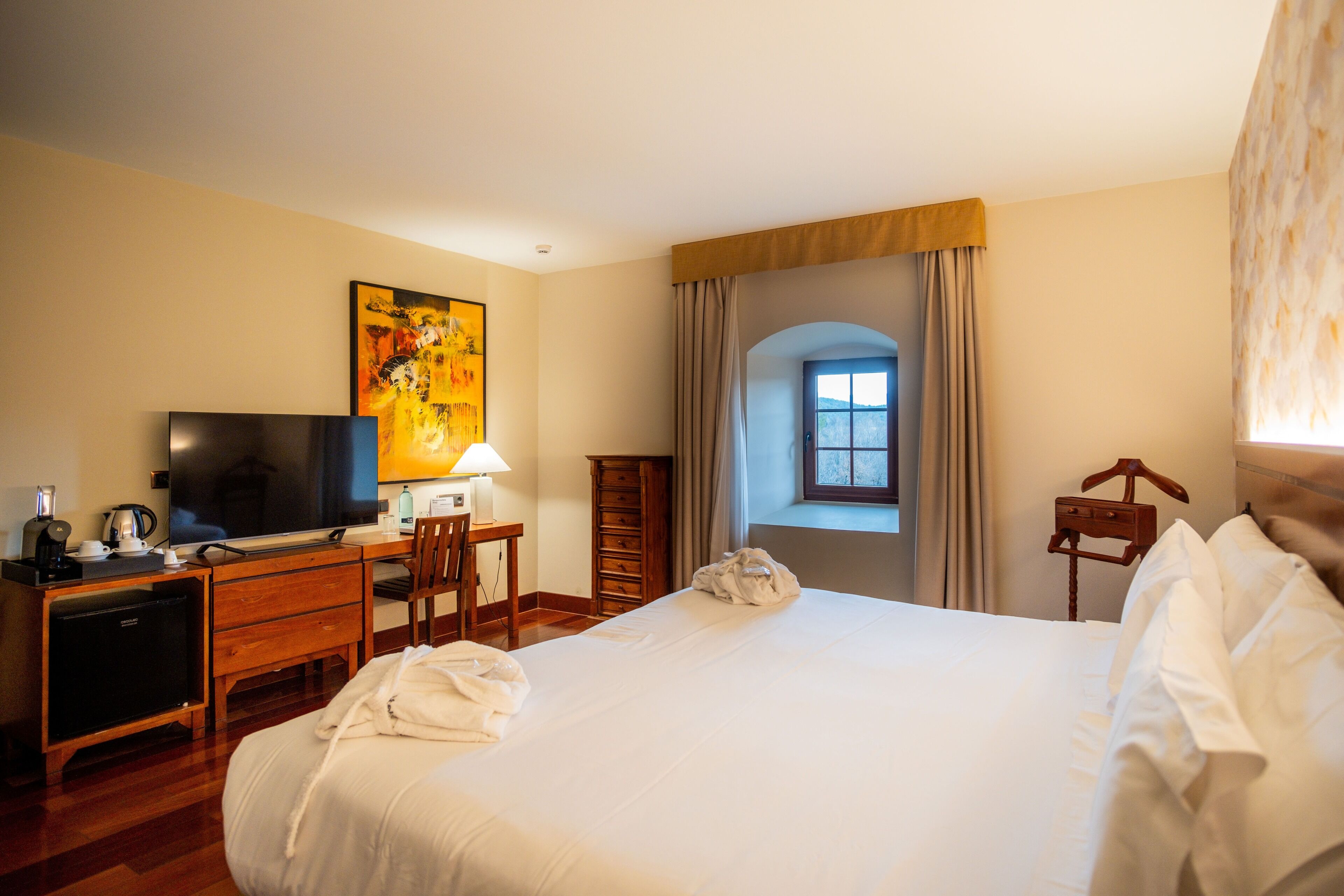 Une chambre d’hôtel avec un lit, un bureau avec une télévision, une lampe et une fenêtre avec des rideaux.