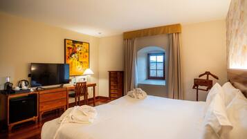 Habitación de hotel con cama, escritorio, televisor, lámpara y ventana con cortinas.