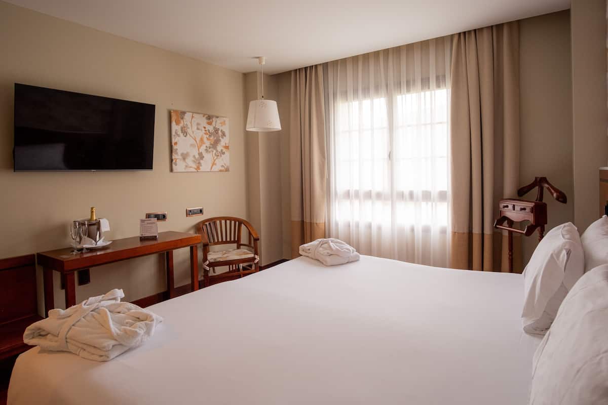 Quarto de hotel com uma cama grande, uma escrivaninha com cadeira, uma televisão e uma janela com cortinas.