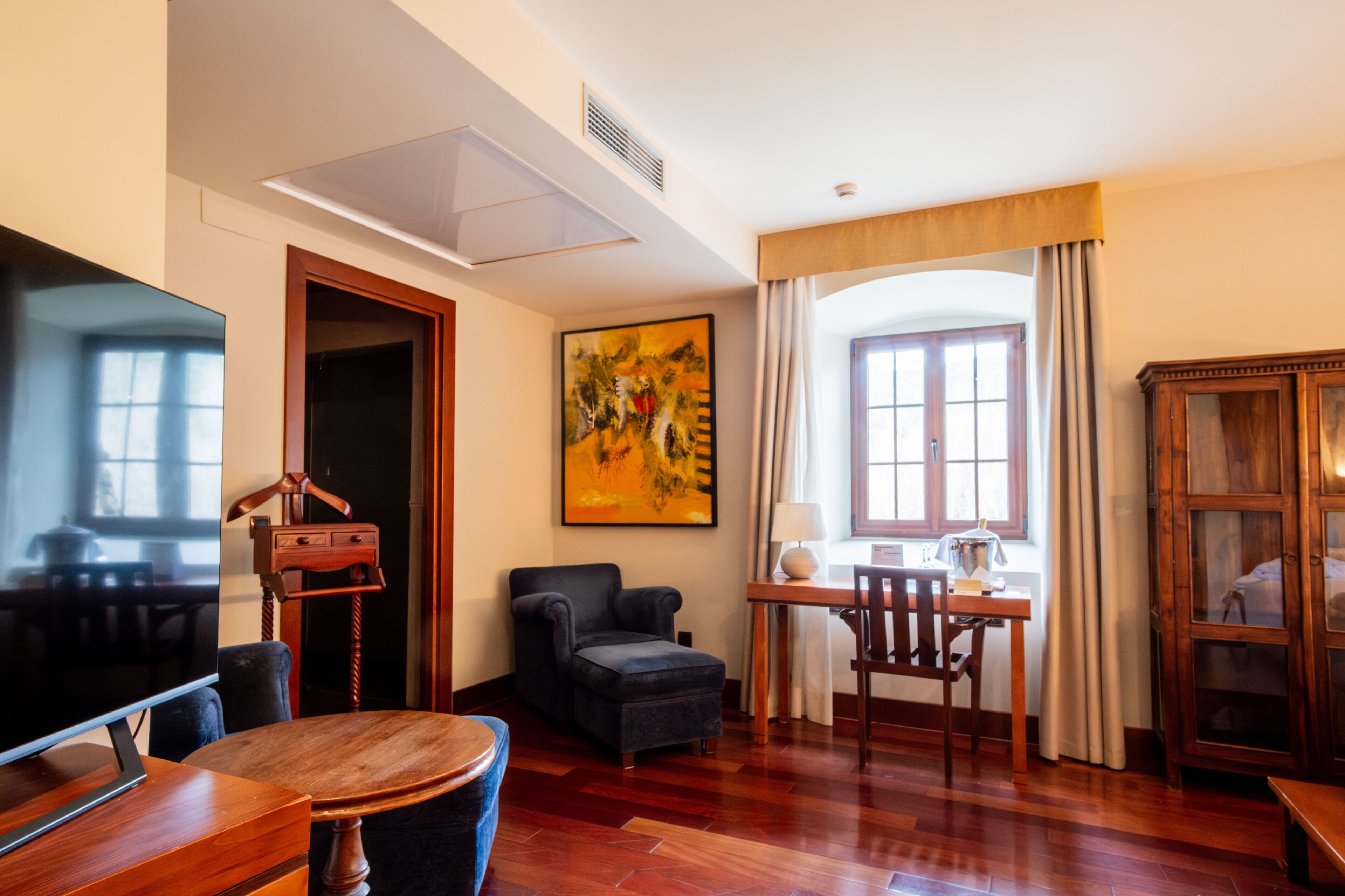 A hotel room with a wooden floor, a flat-screen TV, a dining table, a chair, a sofa, a cabinet, and a painting on the wall.